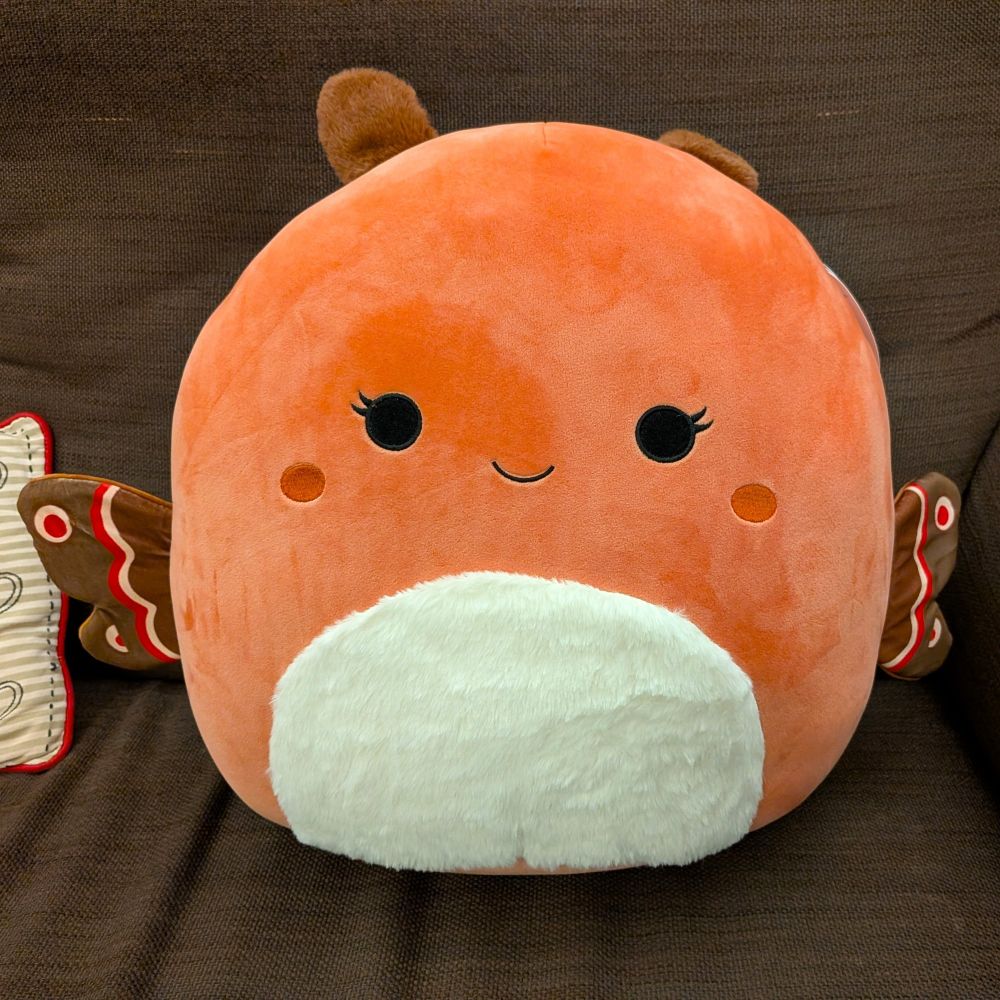 The same moth plushie sitting on a sofa. It's got a big soft round body, a smiling face, two tiny antennas, and two tiny moth wings. Friggin adorable.