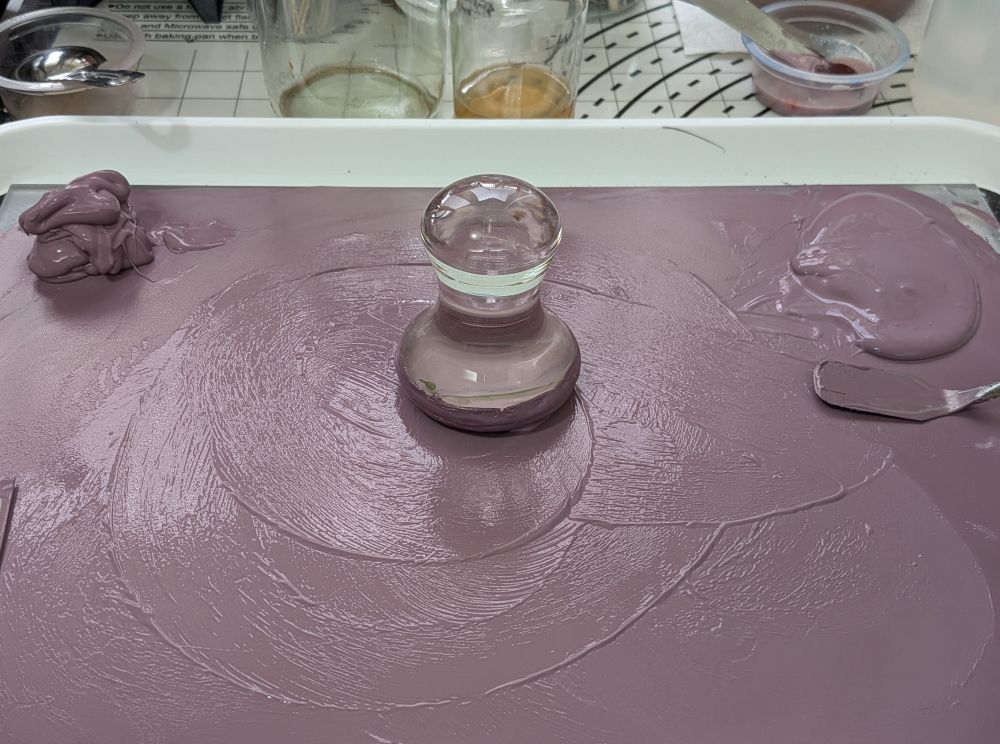 Paint-making in progress. A glass plate is covered in a thin slick of opaque violet watercolour paint, being made by combining pigment powders and a liquid binder. In the middle is a glass mulling tool, which looks a bit like an upside down glass mushroom with a flat top. It's used to smoosh a small amount of paint at a time, pushing it between the surfaces of the plate and muller to thoroughly combine pigment and binder. In the top right corner is a pool of paint waiting to be mulled. Top left is a glossy heap of gloopy fully mulled paint. 