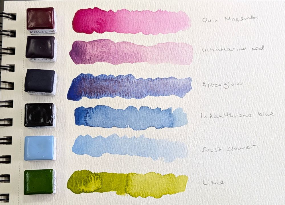 Six half pans of watercolour paint, arranged in a column next to a swatch of each colour. They're labelled quinacridone magenta, ultramarine red, afterglow, indanthrene blue, frost flower, and lime. Two are pink, one is purple, two are blue, and one is green.