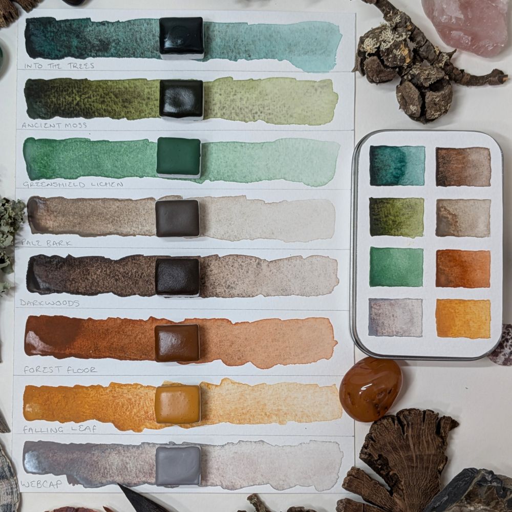 An 8 colour half pan handmade watercolour set. There's a swatch card showing a strip of each colour, and an open pan of each resting on the swatch. There's also a swatched square tin to one side. The colours are granulating and semi opaque mixed pigment colours, mostly in greens and browns because it's a woodland themed set.
