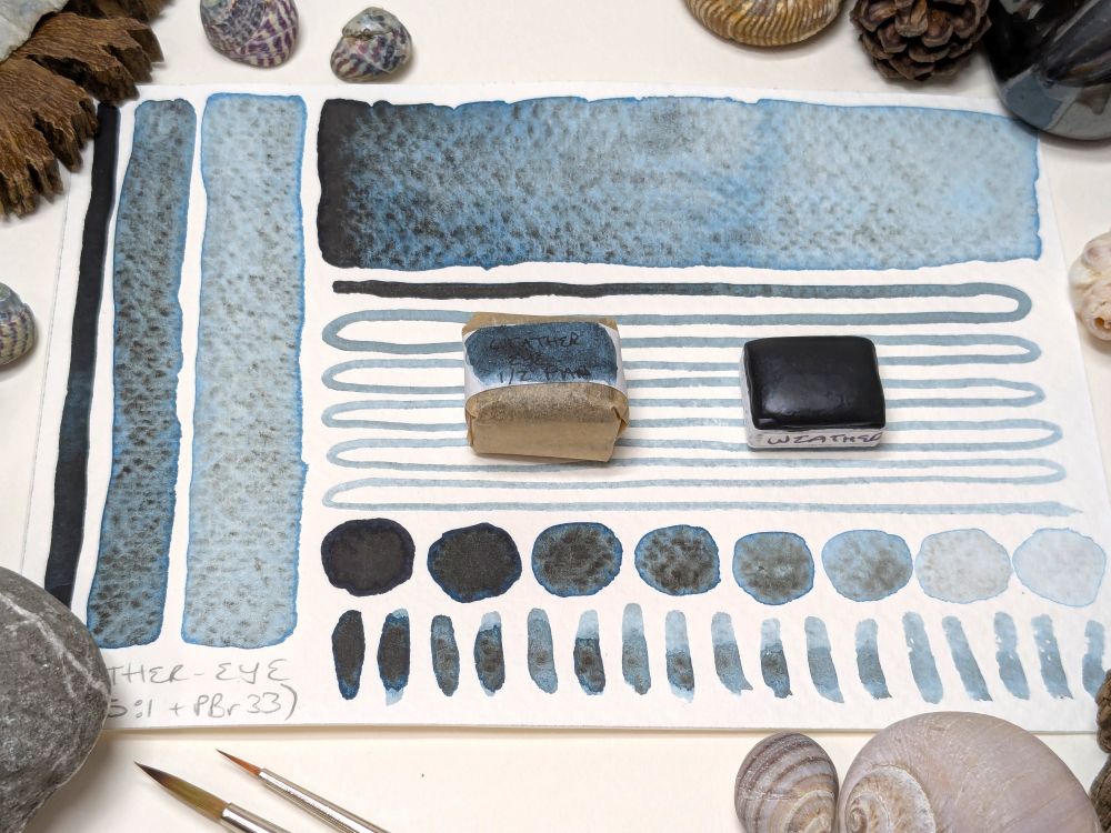 Promotional pic for handmade blackish blue watercolour paint with the blue coming from phthalo blue red shade PB15:1 and the dark granulation coming from chromite PBr33. Two pans are sitting on a postcard swatch that has stripes, lines, and circles showing how the paint looks at different dilutions with water. There's shells, pebbles etc dotted around the edges to pretty things up.