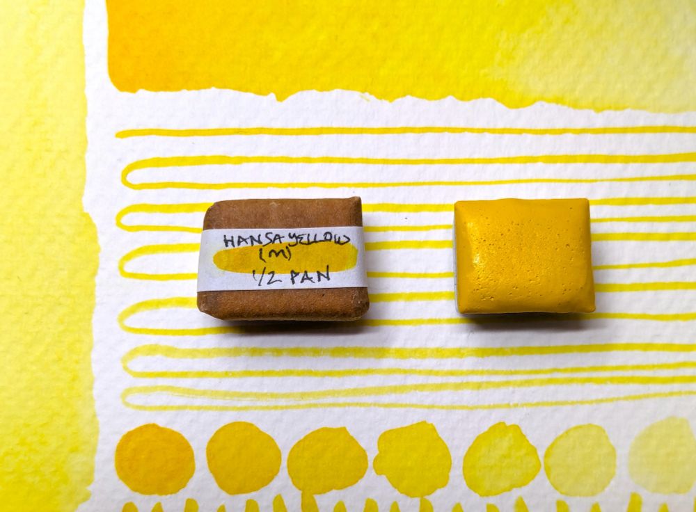 Two pans of Hansa yellow medium PY74 watercolour paint - one wrapped and labelled, one open - sitting on a postcard swatch showing how the paint looks at different dilutions with water.
