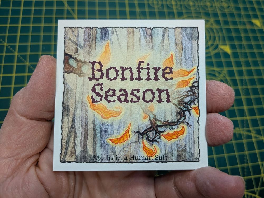 My hand holding the cover card of the stack zine. It has the words 'Bonfire season' over a woodland scene with dark tree trunks and a lichen covered branch surrounded by falling leaves like glowing embers.