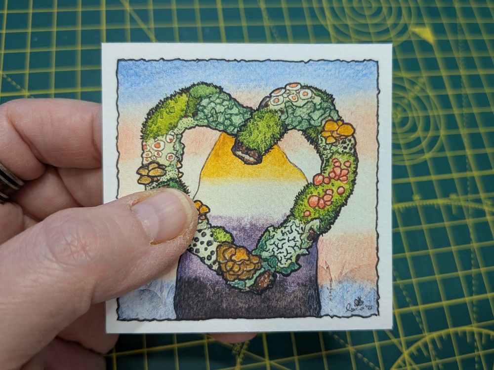 My hand holding up an artwork square from the stack zine. It has the same image as the quoted post - a heart made out of lichen covered branches, the silhouette of a standing stone in non-binary pride colours, and a background sky in trans pride colours.