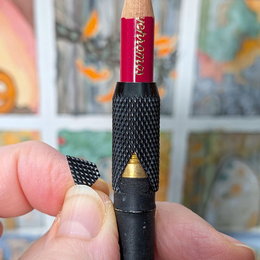 My hand holding up a screw collar pencil extender with a small triangular piece of the metal collar broken off. The collar and most of the barrel are black, but the broken areas reveals the brassy colour of the screw thread. In the background are blurred out autumnal drawings.