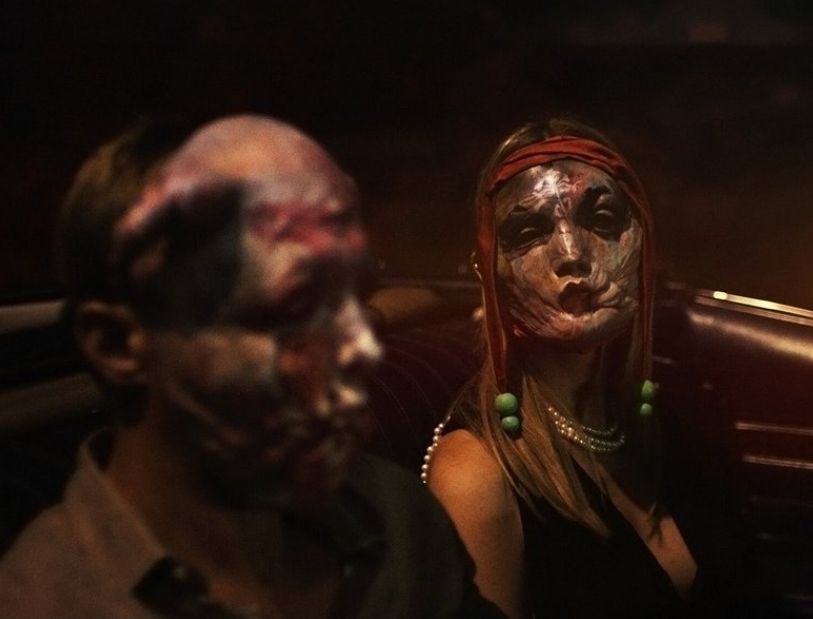 Scene from the Brandon Cronenberg film Infinity pool. Characters played by Alexander Skarsgard and Mia Goth are wearing creepy masks while being driven in a convertible. 