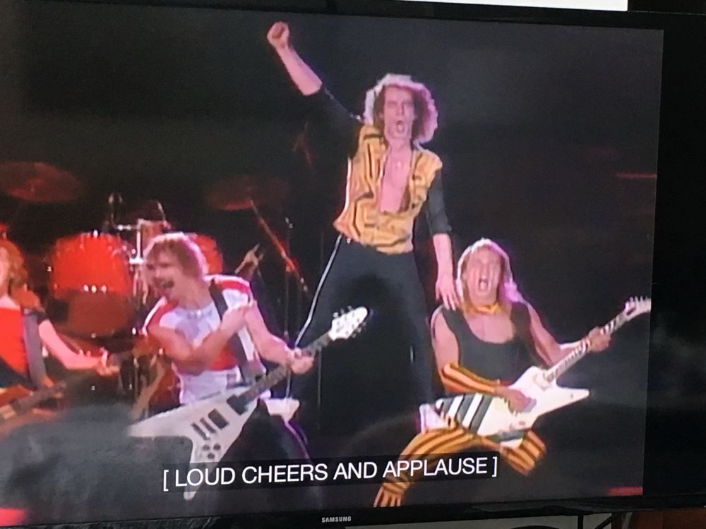 Scorpions playing at the US Festival. Kalisz Meine is raising a fist in the air while standing on the thighs of Rudolph Schenker and Matthias Jabs as they play their guitars. The closed captioning reads (LOUD CHEERS AND APPLAUSE)