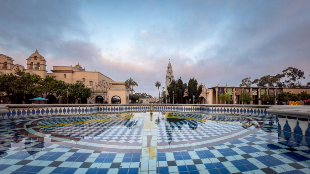 A tall Spanish Colonial tower rises above surrounding buildings in early morning light. The structure is silhouetted against a sky of pastel pink, orange, and blue with soft layered clouds. Additional domes and tree outlines appear on the left, with the warmest sunlight touching the lower rooftops.