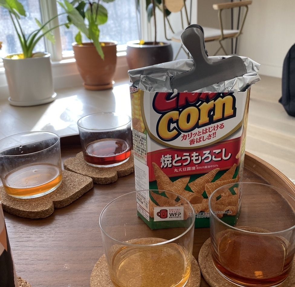 Two sets of two lowball glasses filled with a small amount of a light amber liquid, and a deep mahogany liquid, on a wooden tray. The coasters are shaped like toast. Next to them is a bag of Japanese corn snacks and there are houseplants in the background. 