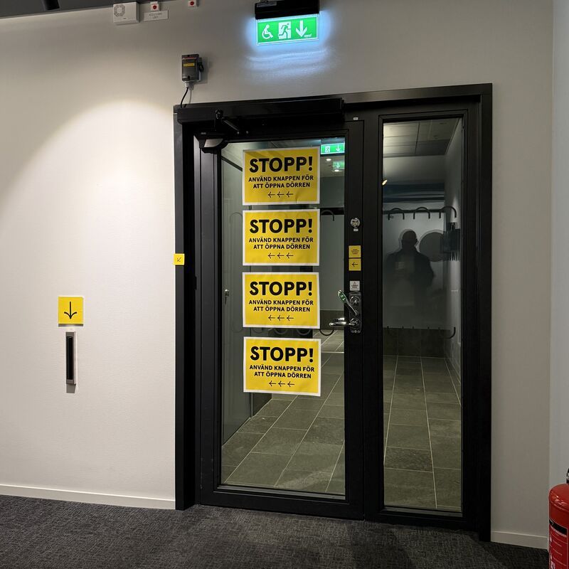 A door with four big yellow signs printed out and taped to it that says stop, press the button to open the door. They point to the left, where another arrow points down towards a button.