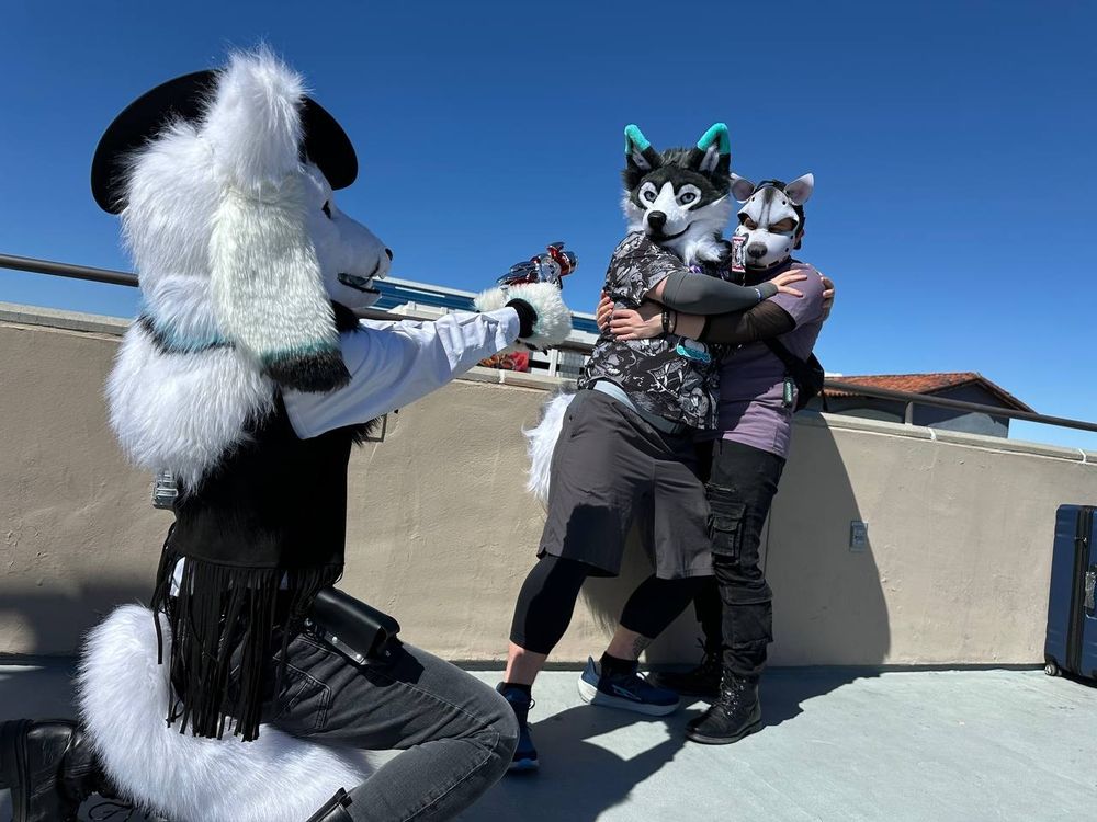 Axen Foxgoat dressed as a space cowboy on a sunny balcony squats in the foreground, aiming his raygun at a frightened Ceeps Husky and Sinhto Coyote, holding each other for safety as they stare into the barrel of the raygun. 