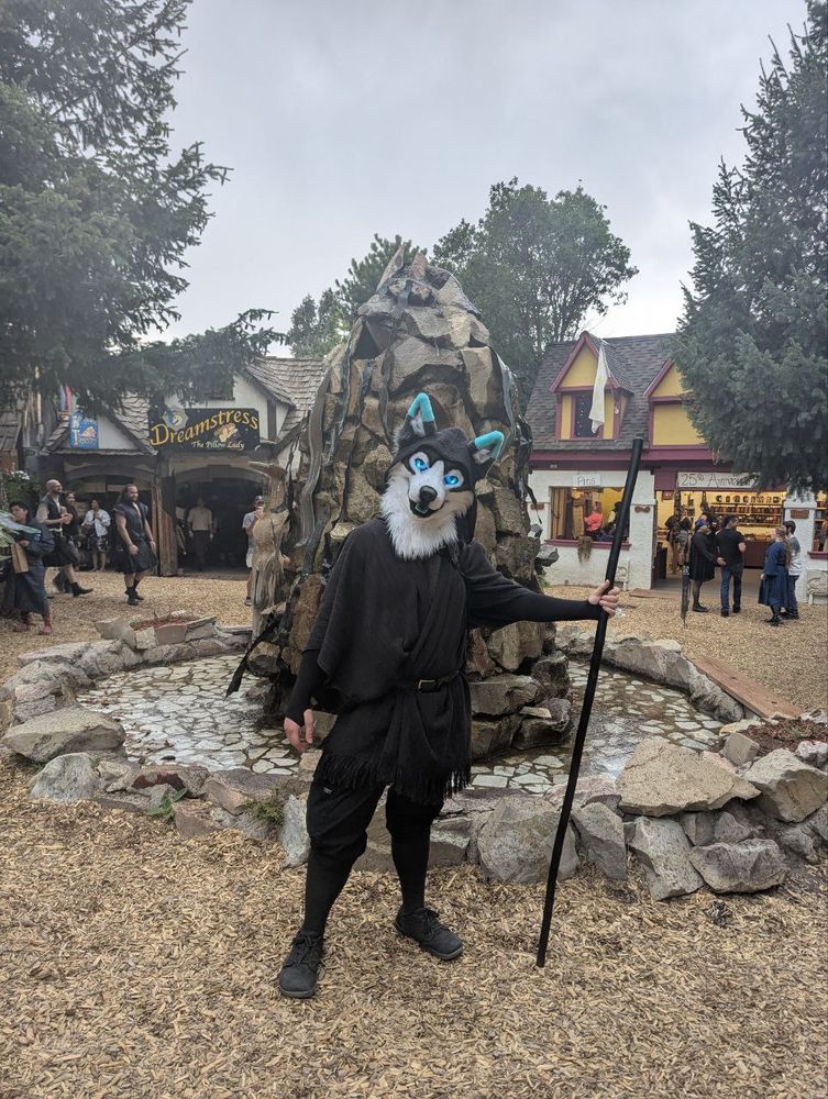 Ceeps Husky standing in front of a fountain at the Colorado Renaissance Festival in his hooded wanderer outfit, head cocked and peering a glowing icy blue gaze toward the camera. 