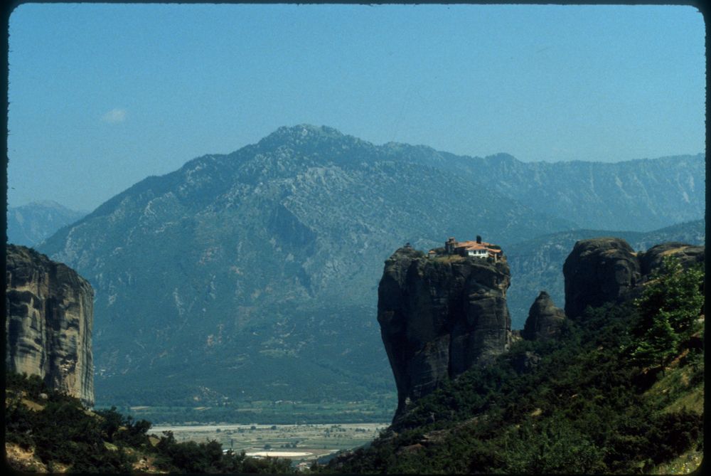 A 35mm slide of a landscape with a large mountain in the background, a green valley in the foreground, and a clay-roofed, white-walled house at the very top of a large column of stone just right of centre. The house on the stone pillar is a monastery in Meteora, Greece.