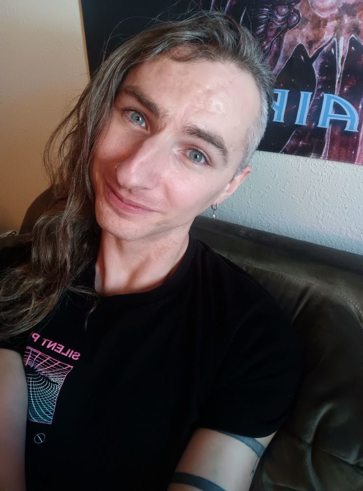 Anna Lee, white queer/trans person with long wavy brown/grey hair shaved on the sides and blue eyes, with a silver nose ring, wearing a black silent planet tee, sitting on the couch and smiling gently