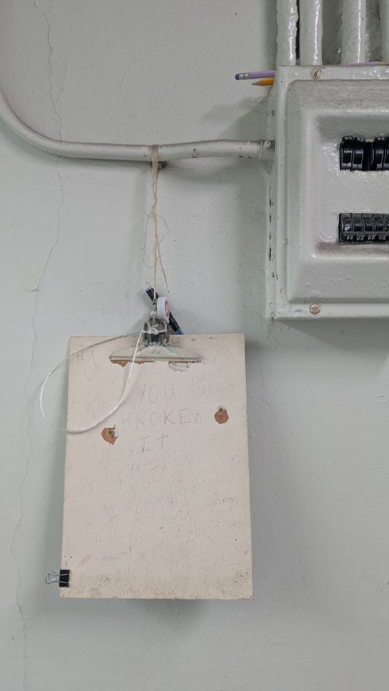 Clipboard hanging by frayed twine from a cable feeding into a breaker box, with a pen tied to it.  The clipboard itself is old, with chipped white paint that doesn't match the blue of the wall behind it, and people have scratched things into it, but the only legible scratched message is a large, central, all-caps "YOU BROKE IT" that looks almost red against the white paint.  A single binder clip is attached to the lower left side of the clipboard.
