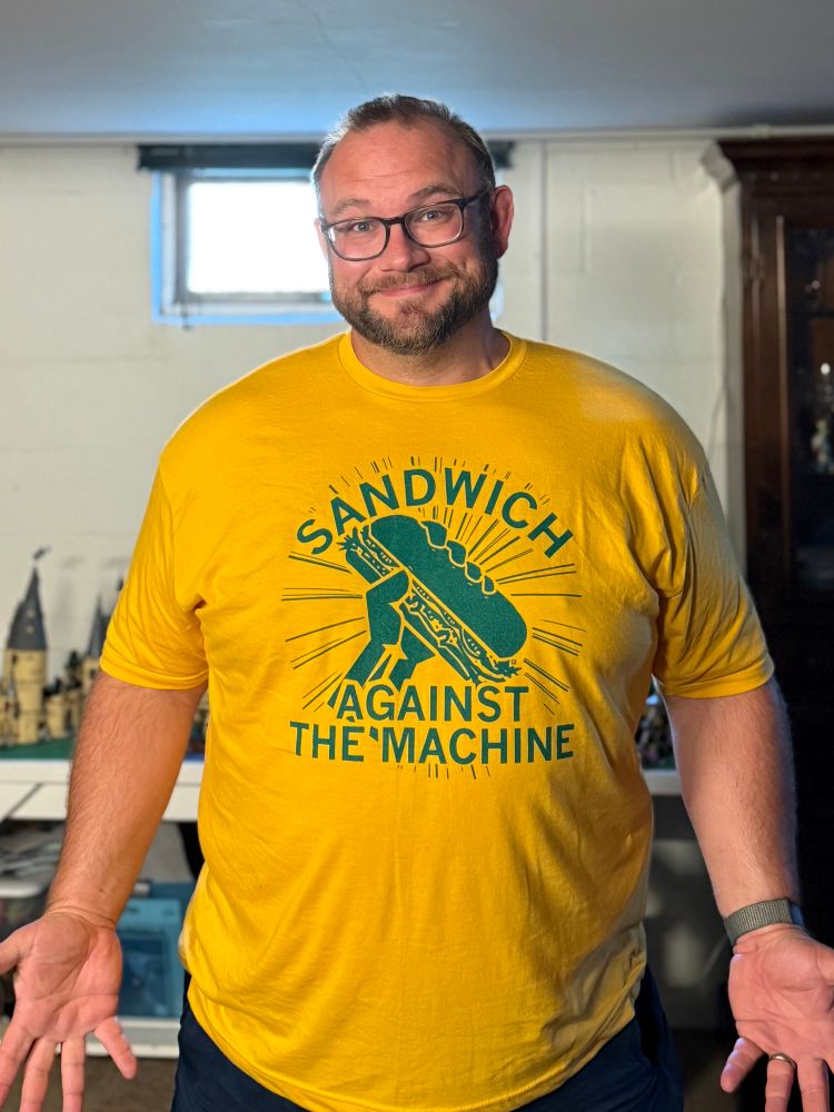 A robust, bearded white 40-something male stands in a yellow 3XL shirt. The shirt reads SANDWICH AGAINST THE MACHINE.