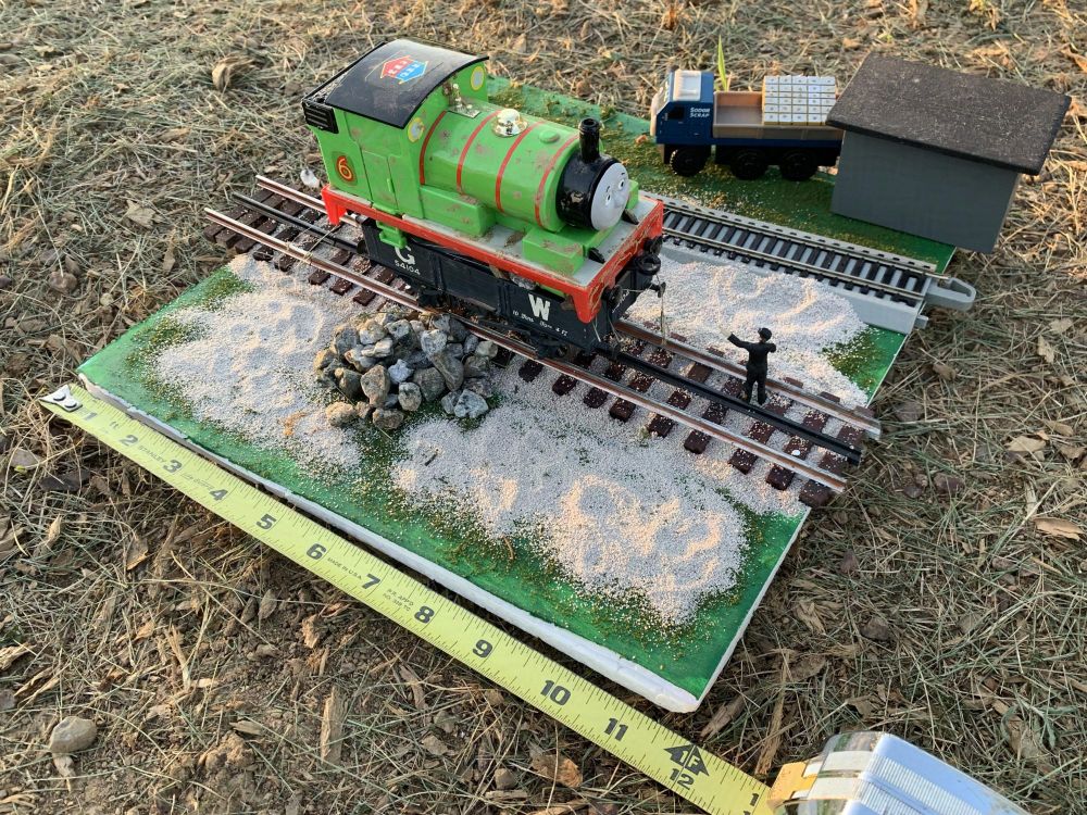 Full color photograph of a small light green saddle tank steam locomotive with a red chassis, four driving wheels, and red stripes on his boiler, with a yellow no. '6' outlined in red behind his cab and a black roof on top with yellow circle windows for the crew to see through. He is perched atop a black open wagon sitting on the railway tracks lettered "G W" for Great Western, as well as labels "54101" and "10 Tons". The locomotive is Percy from the Thomas the Tank Engine/Railway Series franchise, as he has a sad expression and is covered in dirt. Beside the wagon and next to the tracks is a pile of stones spilled from it. In front of the truck is a black suited gentleman, the Fat Controller/Sir Topham Hatt, scolding and pointing at Percy for his predicament. The camera is pulled out to show it is an approximately 12 inch by 12 inch diorama display in O gauge with a tape measurer showing its dimensions.