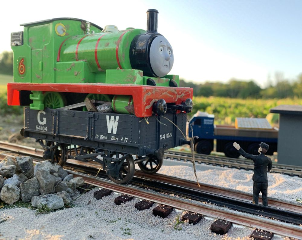Full color photograph of a small light green saddle tank steam locomotive with a red chassis, four driving wheels, and red stripes on his boiler, with a yellow no. '6' outlined in red behind his cab and a black roof on top with yellow circle windows for the crew to see through. He is perched atop a black open wagon sitting on the railway tracks lettered "G W" for Great Western, as well as labels "54101" and "10 Tons". The locomotive is Percy from the Thomas the Tank Engine/Railway Series franchise, as he has a sad expression and is covered in dirt. Beside the wagon and next to the tracks is a pile of stones spilled from it. In front of the truck is a black suited gentleman, the Fat Controller/Sir Topham Hatt, scolding and pointing at Percy for his predicament. In the background is a small blue and gray lorry/truck next to a small warehouse. The sky is blue and the background shows many green hills and trees. The scene is based off the story 'Percy's Predicament' from the second season of the TV series, and the fourth story in book no. 16 "Branch Line Engines."