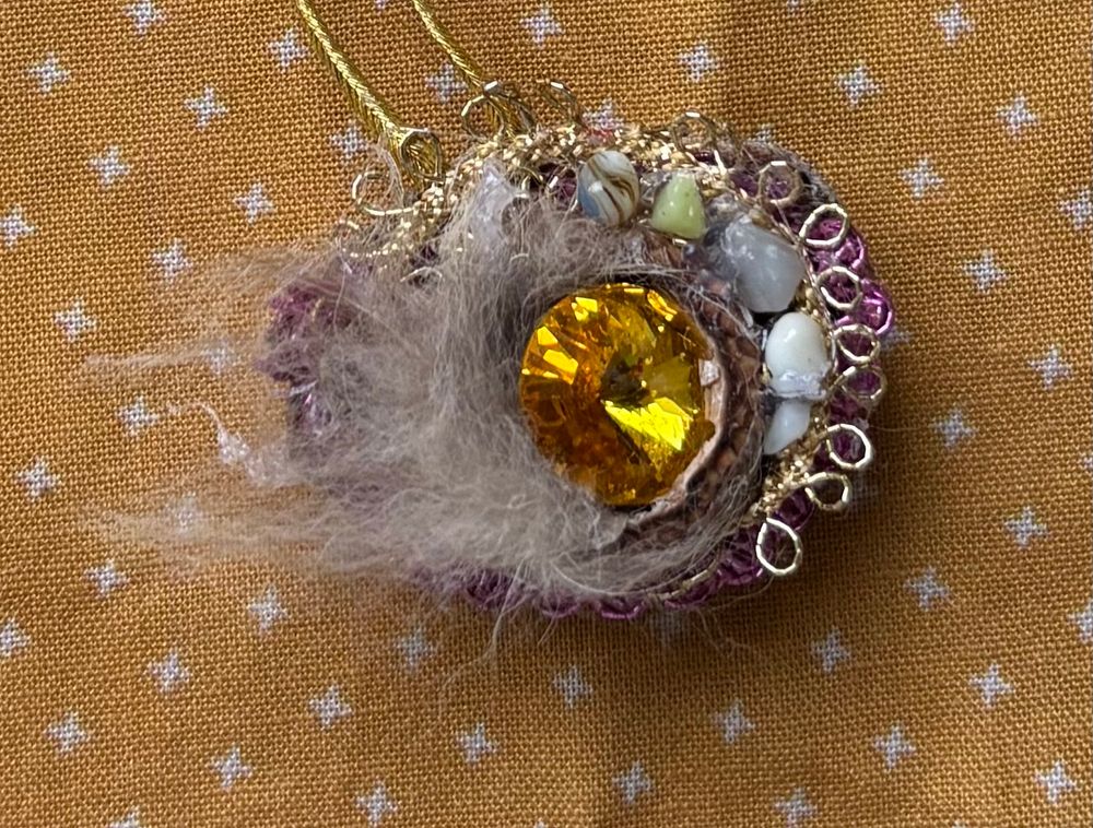 A gold cabichon is nestled inside  an acorn cap inside a glazed milk weed pod. Tiny stones and fabric edging along the edge of the pod. Tuft of wolf fur extends from left side when the pod is viewed horizontally.