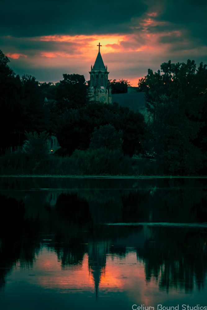 A sunset behind a church steeple, with the brightest spot being directly behind the cross. The church is surround by trees. The scene is then reflected off the lake. "As Above, So Below" or something like that