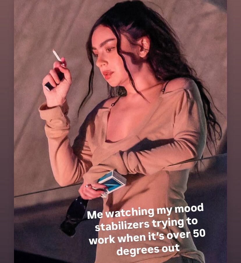 Charli XCX looking intensely over a music video filming - smoking a ciggy and drinking a beer and looking very stressed. Text over picture says ‘Me watching my mood stabilizers work when it’s over 50 degrees out’