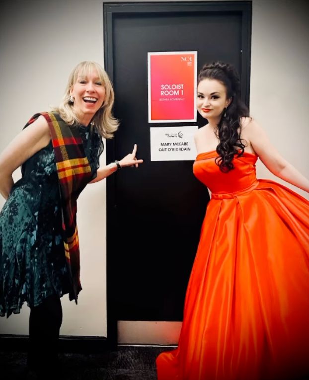 two singers in fancy gúnas pointing at their names on a dressing room door backstage at the National Concert Hall in Dublin, Ireland. One singer is wearing a gorgeous fiery red silk ballgown and has extremely elegant long curled opera star hair. The other singer is in a floaty green dress with a yellow and red tartan sash. They are both wearing sparkly earrings for the night that's in it