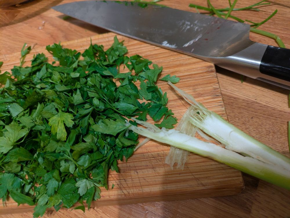 Image of chopped parsley and chef's knife on a wooden chopping board with a stalk of fennel that's clearly been chewed at one end
