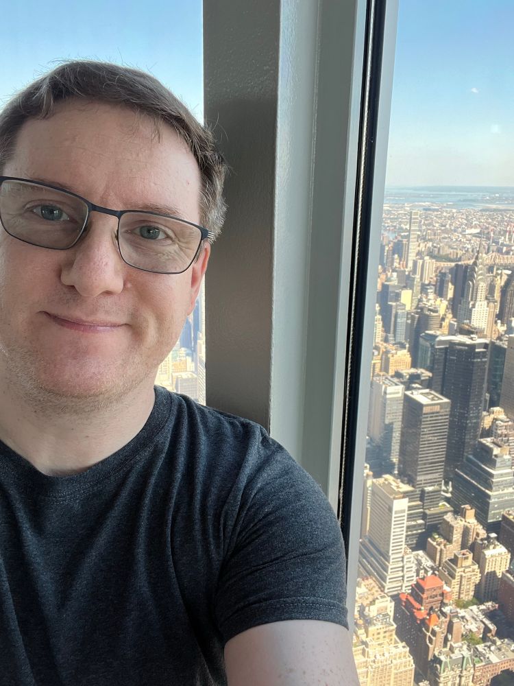 Some dude in glasses smiling at the camera. To his left is a cityscape seen from a very tall building. 