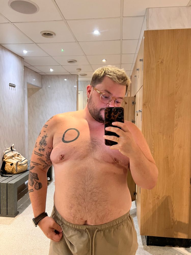 Ocean is taking a shirtless selfie in front of a mirror against an empty locker room
