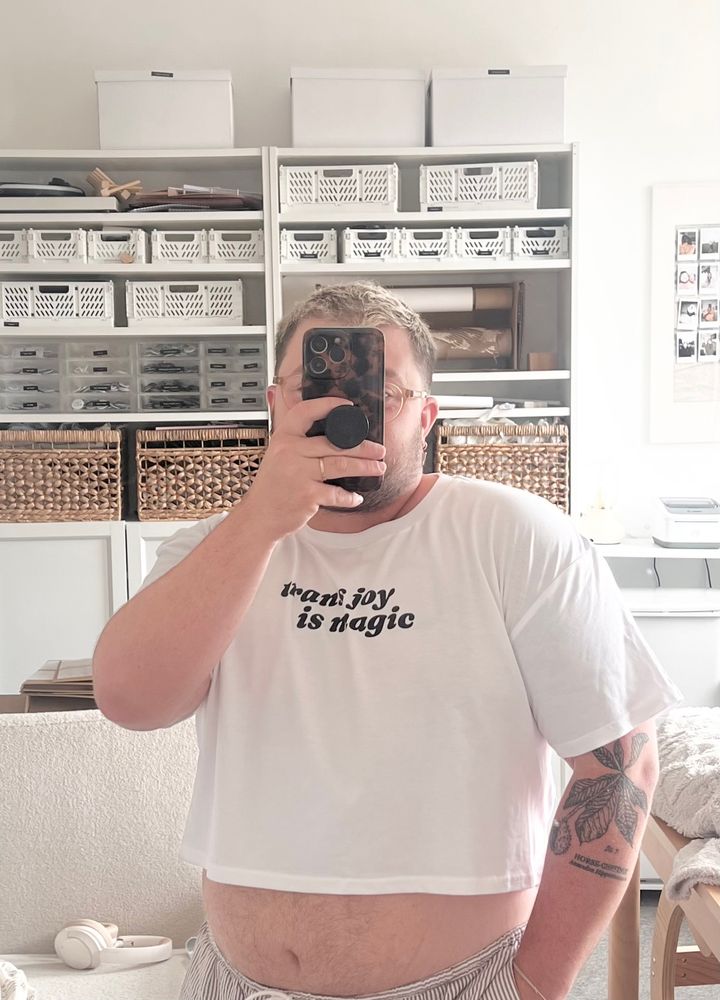 Ocean is wearing a white cropped top that says “trans joy is magic” on it in serif writing. He’s taking a selfie in the mirror 
