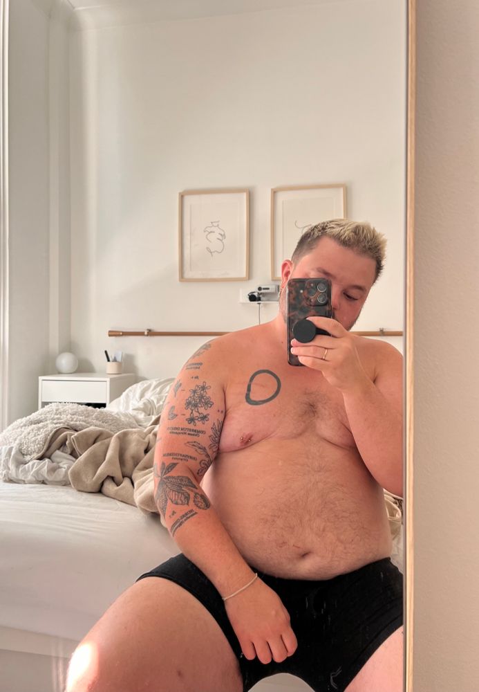 Ocean is sitting at the end of his bed in black boxers. He’s taking a selfie in the mirror against his neutral bedroom in the background