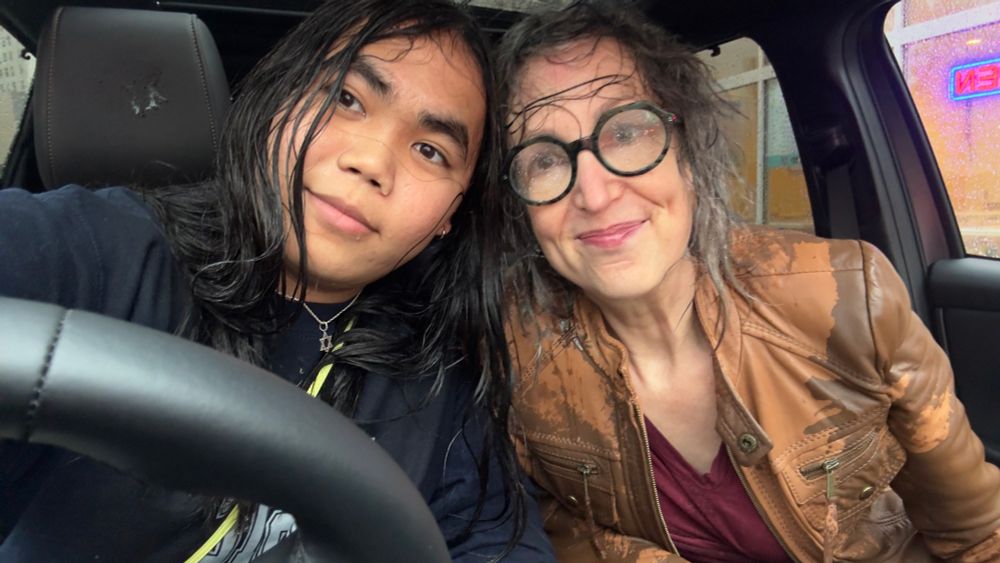 a white mom and asian teen girl lean into each other in the car, faintly smiling, hair plastered to their faces, clothes soaked. mom wears large rain-splashed glasses.