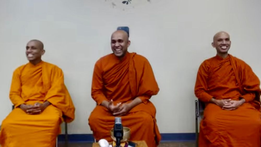 three sri lankan monks sitting side by side in front of the clean white wall of a public library. their robes are three different shades of saffron.
