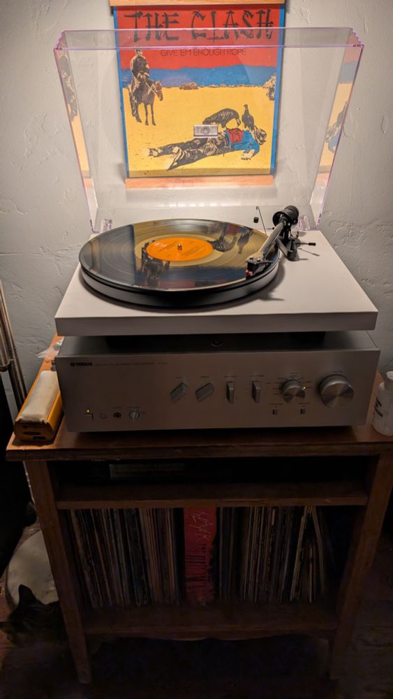 A vinyl album cover on a "now playing" display over a component stereo.