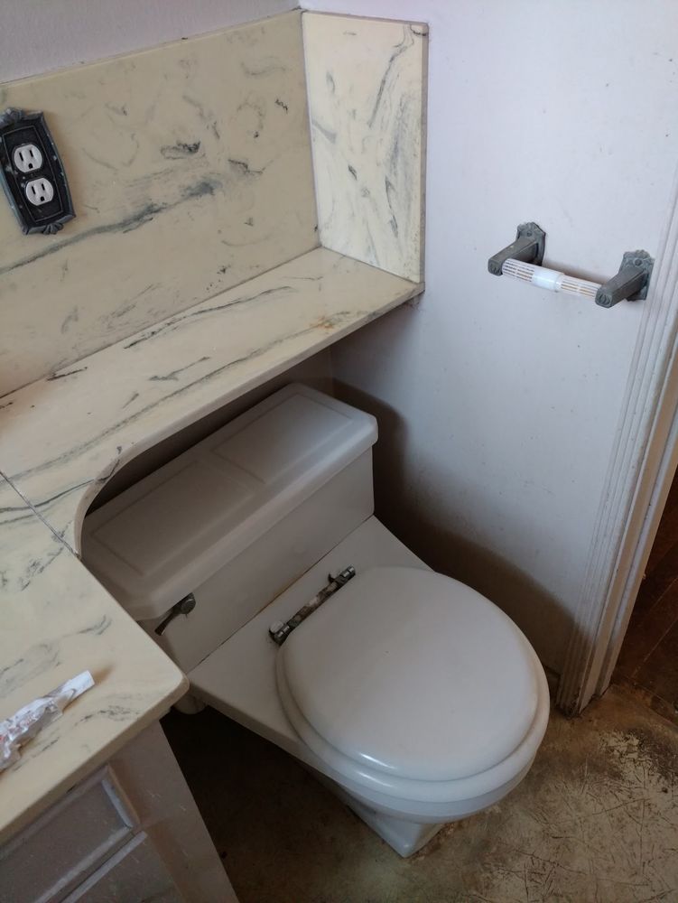 A picture of an older bathroom with a worn linoleum floor and an empty toilet paper holder. 