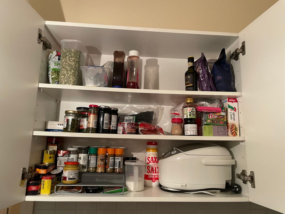 An upper kitchen cupboard is open to show assorted foods and a rice cooker placed much more neatly and accessibly than was previously the case.