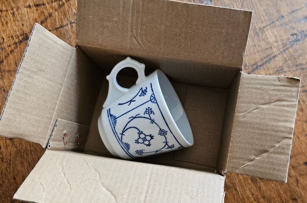 Alte weisse Tasse mit Muster Indisch Blau in einem kleinen Karton auf einem alten Holztisch.