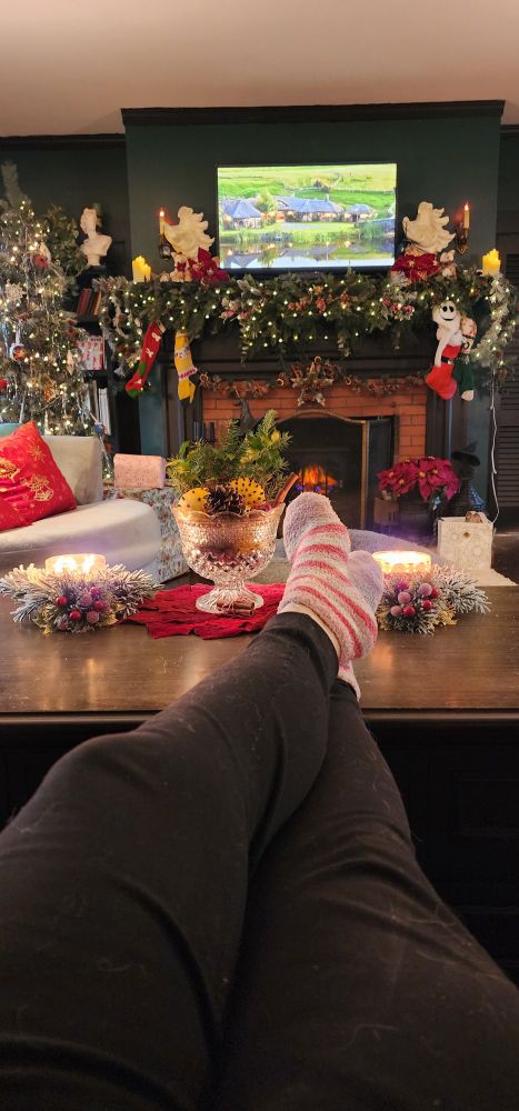 Two feet wearing pink fuzzy socks are  crossed at the ankle on a coffee table.  2 lit candles, a crystal bowl filled with pinecones, pomanders, cinnamon sticks, cranberries, evergreen clippings and Holly clippings also sit on the table.  There is a lit fireplace with a big vase full of poinsettas and a wicker witch to the right of the fireplace and a Christmas tree with white lights, silver tinsel and presents underneath to the left.  Above the fireplace is a garland with various evergreen sprays, lights and berries.  On the mantle is a tv with a little Shire (From LOTR) on, two large poinsetta flowers, two ghosts and candles are on either side of the TV