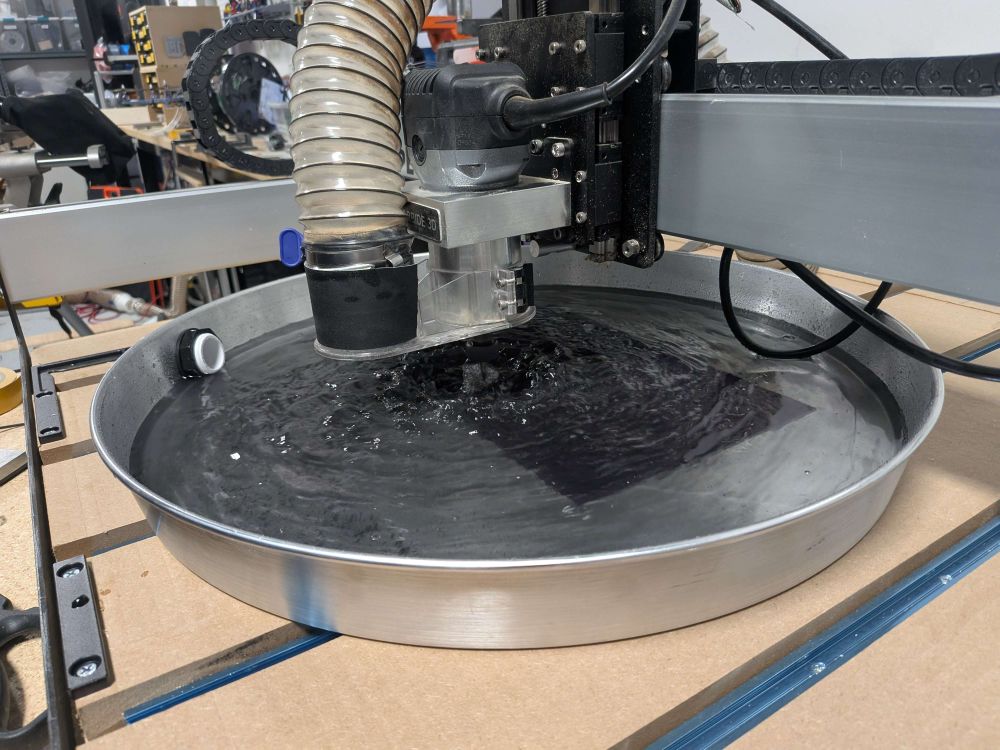 A CNC router (a Shapeoko) milling carbon fibre submerged in a round, metal water bath.