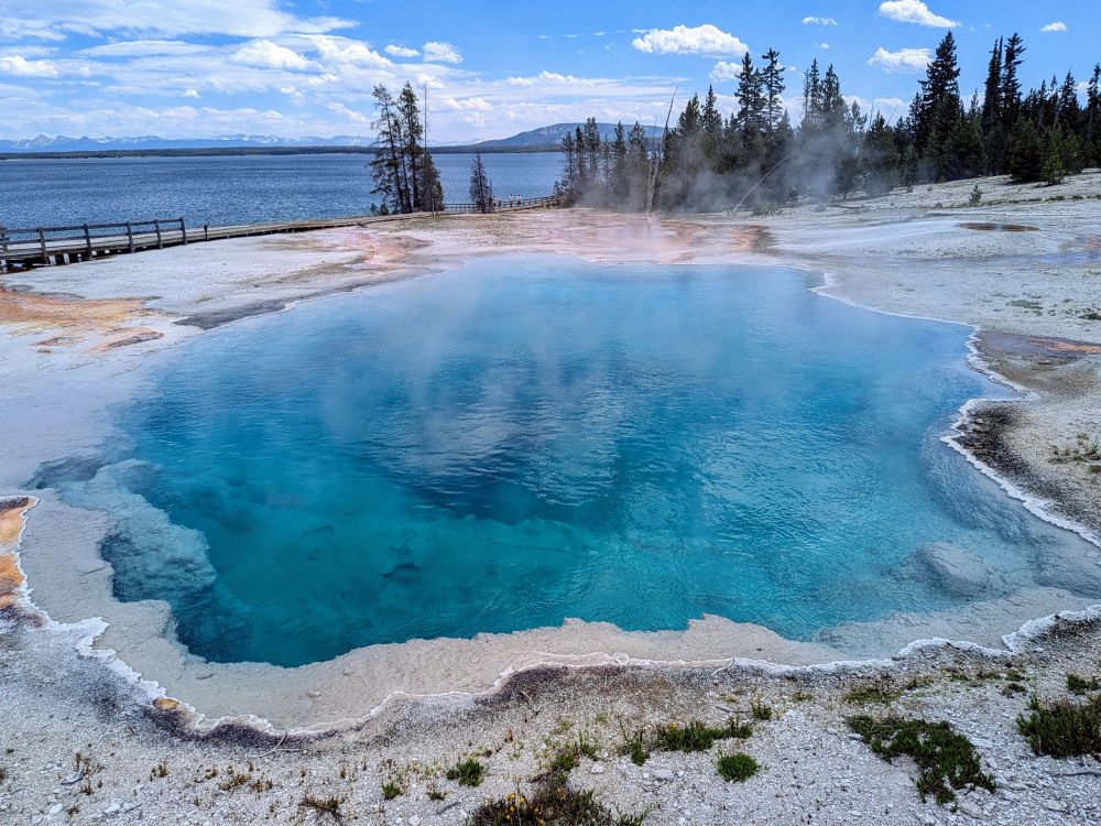 A clear, cerulean blue boiling pool in the middle of a thermal area, steam coming off it's surface, with a lake and distant mountains in the background.