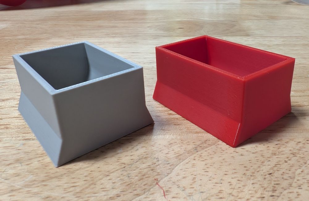 Two 3D prints of a roughly rectangular shape sit on a wooden table. One is gray, one is red, but their quality looks very similar.