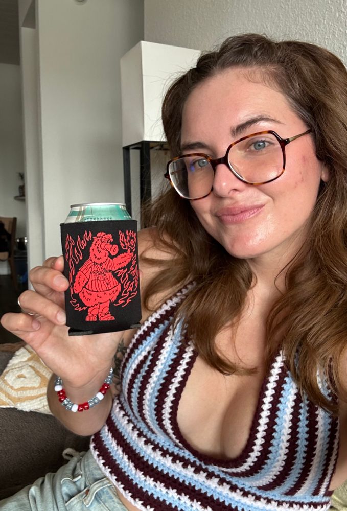 Woman wearing a crocheted maroon, white, blue halter top and jeans. Holding a black and red Phanatic koozie with flames