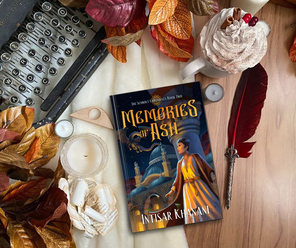 1. A flatlay with 3D mockup of the hardcover of Memories of Ash. The cover shows a young biracial woman with Japanese Arab features. She wears a long embroidered dress belted at the waist, and an Arab-style open-fronted robe. Behind her rise the arched domes of an Ottoman-style school of magic. In the sky, a dragon in flight looks down at her. The flatlay contains additional decorative elements as follows, clockwise from the top: autumn leaves; a white mug topped with whipped cream, cinnamon, and berries, an ornate quill pen with a red feather, the book mockup, a paper flower, additional leaves and small candles, and in the top left corner the corner of a vintage-syle typewriter