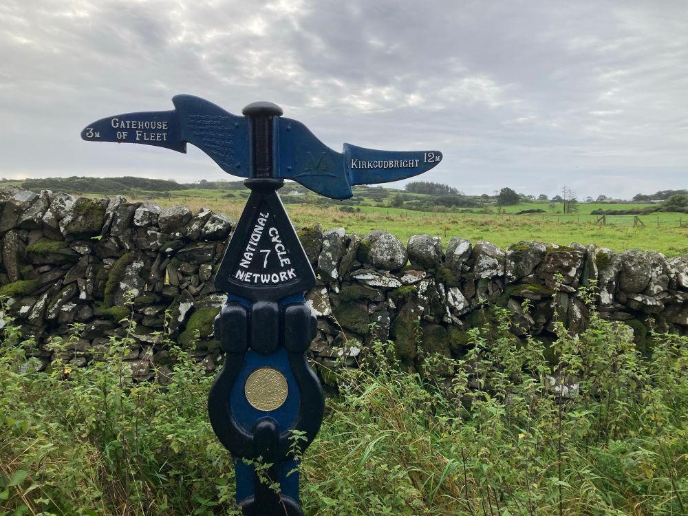 Metal Waymarker for a National Cycle trail. Stone wall behind, then fields. Grey skies.