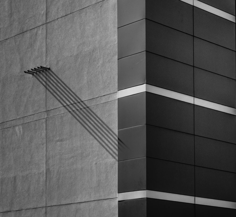 A black-and-white photo focused on the corner of a modern building. On the left, a textured concrete wall is visible. On the right, the building is clad in dark panels. A series of parallel, diagonal shadows from a gutter on the concrete wall stretch across the textured surface.