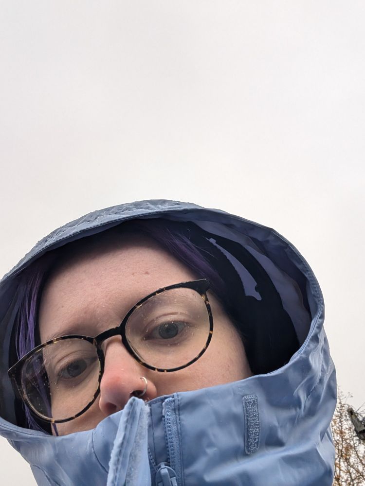 Chloe, a white femme person in a blue waterproof coat with the top done up to cover their mouth. Behind them is endless grey sky. 