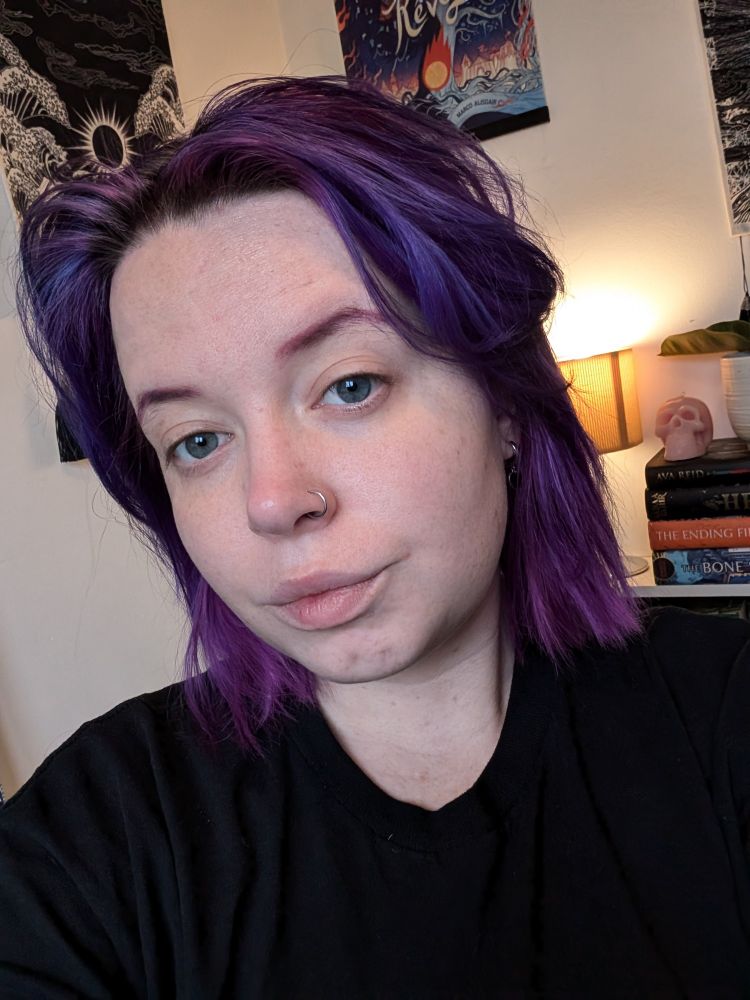 Chloe, a white femme person with purple hair and a nose ring posing in a selfie. Behind them are a number of art prints on the wall, and a bookshelf with a pink skull candle. 
