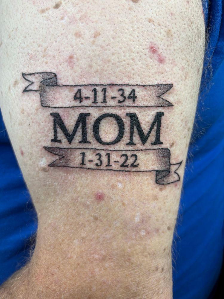 A simple line work tattoo done by cynister designs of the word “mom” with the dates of her birth and passing above and below the text on simple ribbons with minimal shading. 