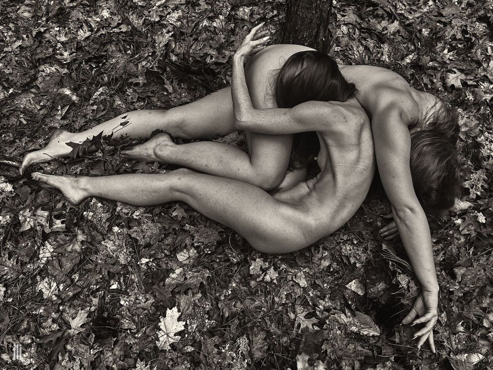 Two nude female art models lie on leaves on the forest floor, partially entangled with one another. Image is monochrome. 
