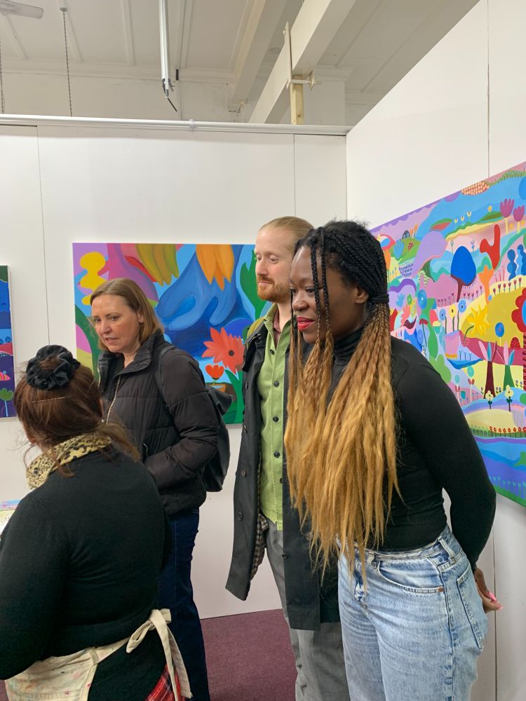 Vicky (centre-behind) and Lewisham councillors listening to Art Hub artist named Mamon speaking about her colourful work which adorns the white walls around them.  